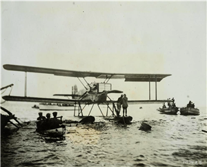 Two people stand on the pontoon of a single prop airplane that is resting on the water. Around them, small wooden boats full of onlookers